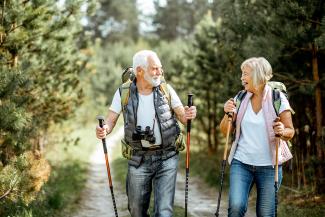 Happy senior couple hiking with trekking sticks and backpacks at the young pine forest.